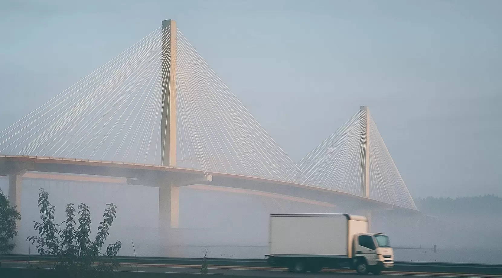 Small box truck in motion beside a foggy cable-stayed Port Mann bridge on a grey morning.