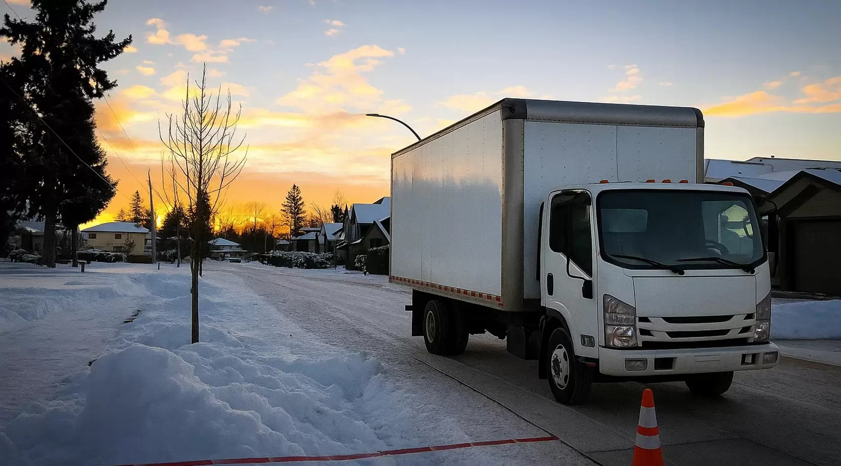 White box truck parked on a snowy residential street at sunrise, marked by a safety cone and pink-gold clouds.