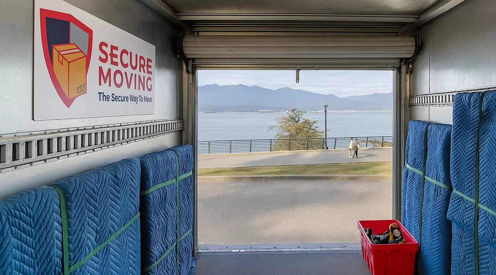 Inside organized truck, stacked pads and strapped furniture. Through open door, Seawalk and mountains show local context. Secure Moving logo as magnetic placard and glove patch. Lighting, clean rails, order.