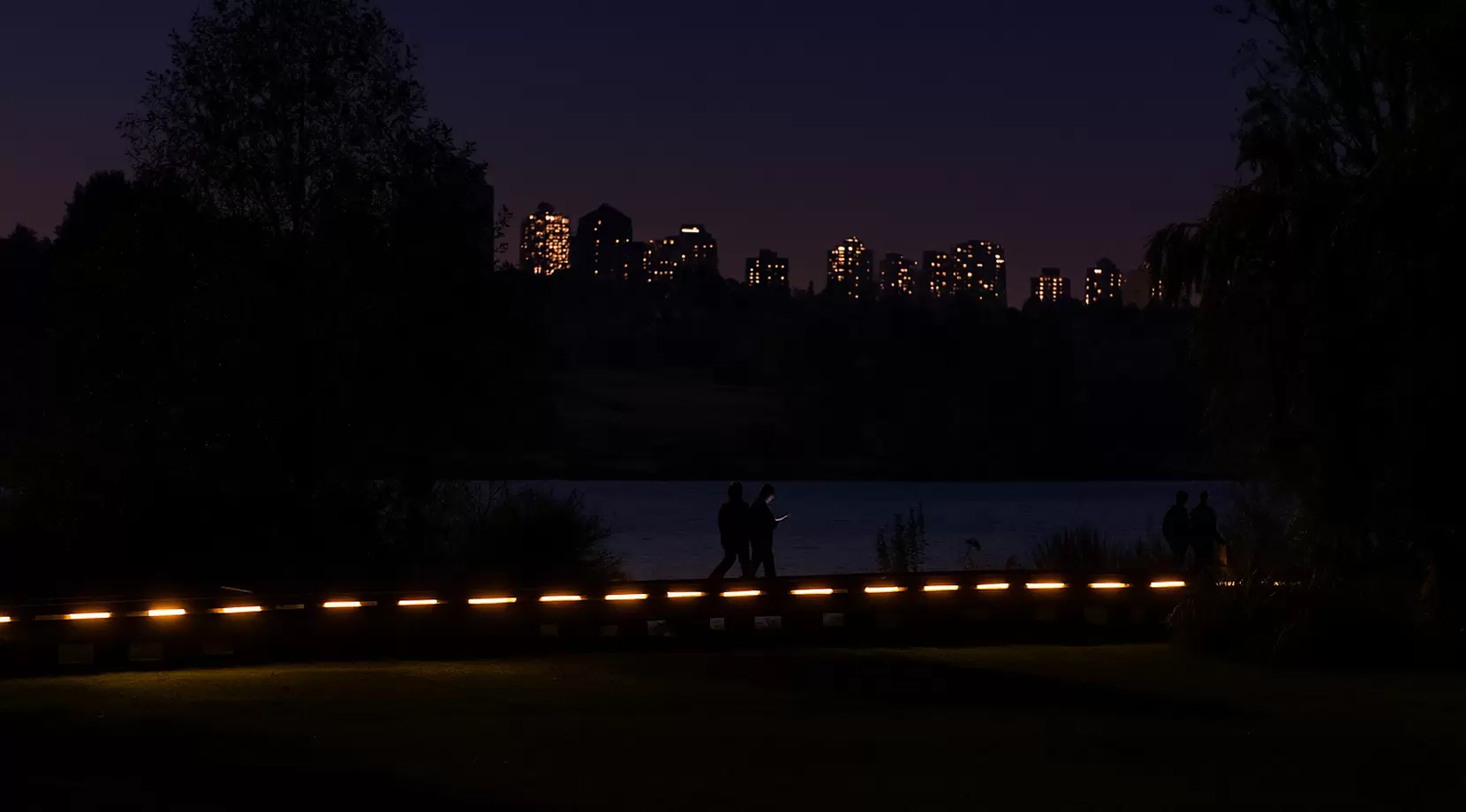 Park walkway after sunset with path lights. A family walks; a parent checks a phone. 
