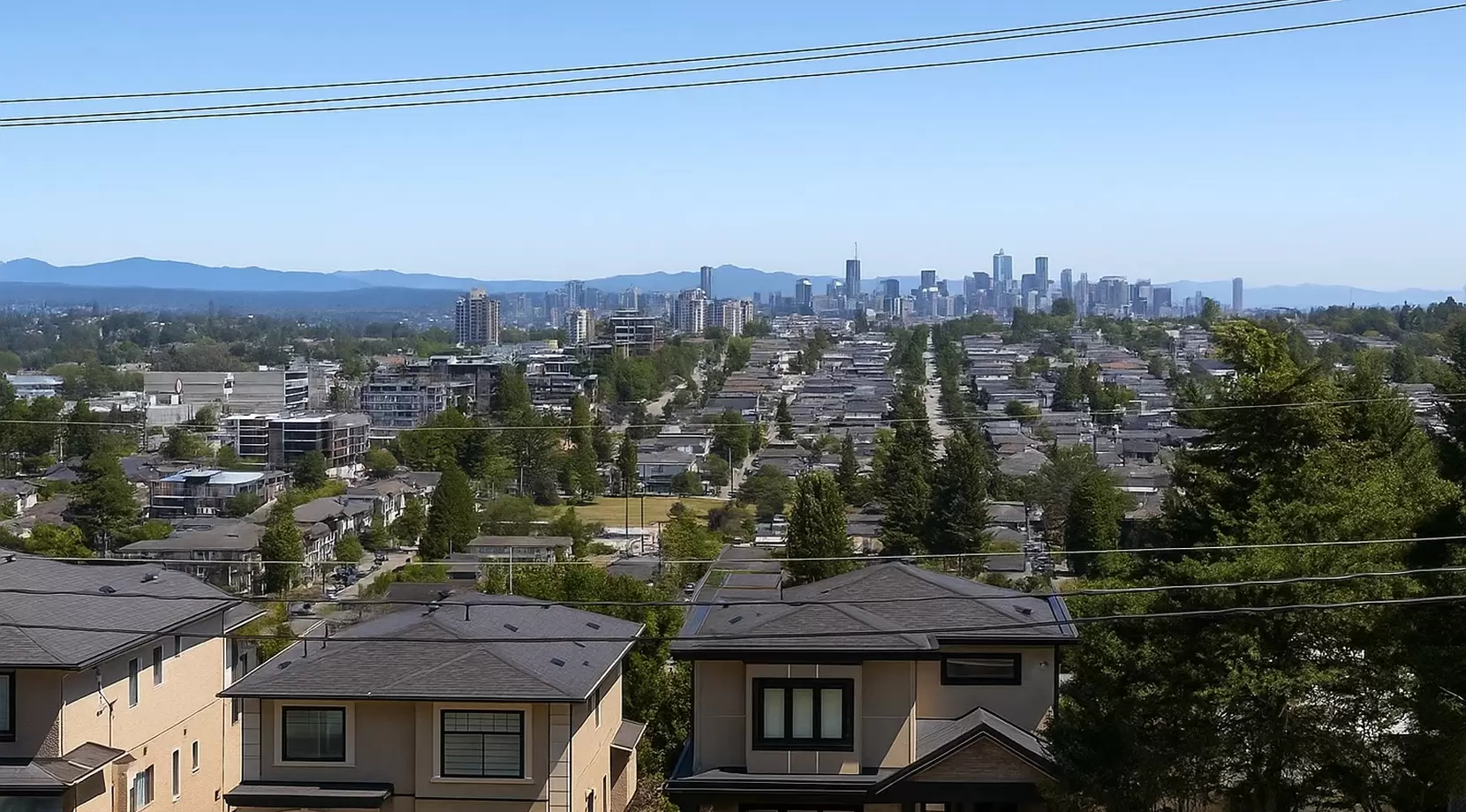 Long view from the hill over rows of low-rise condos toward a distant band of skyscrapers on the horizon. Single-family homes and utility lines frame the clear, blue-sky panorama.