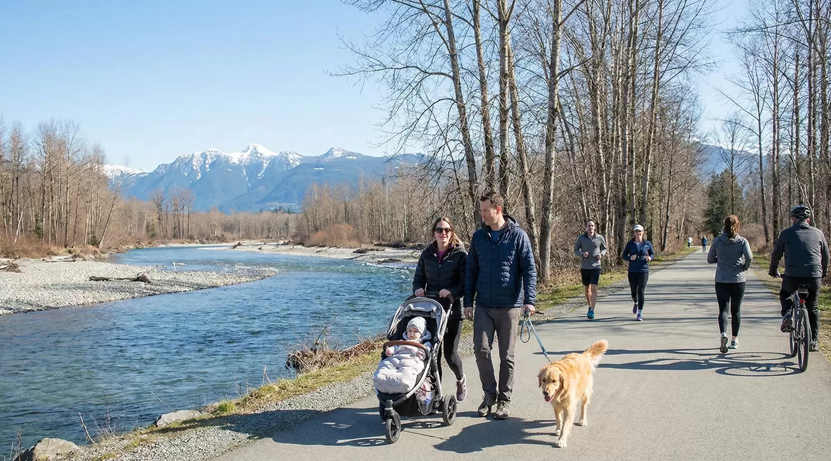 Riverside trail with strollers, joggers, dog walkers, and mountain views, highlighting walkability in Chilliwack and pet-friendly living in Chilliwack.